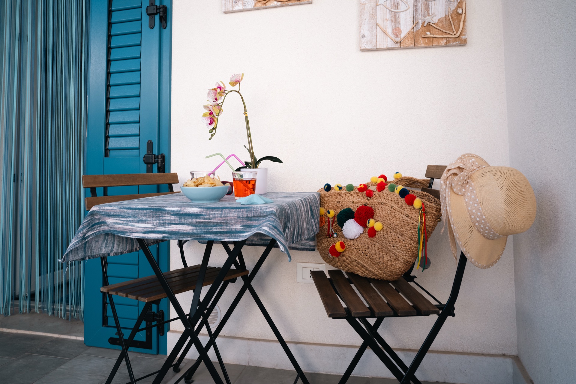Tavolino bistrot con orchidea, spritz, borsa di paglia con pom-pom e cappello da sole